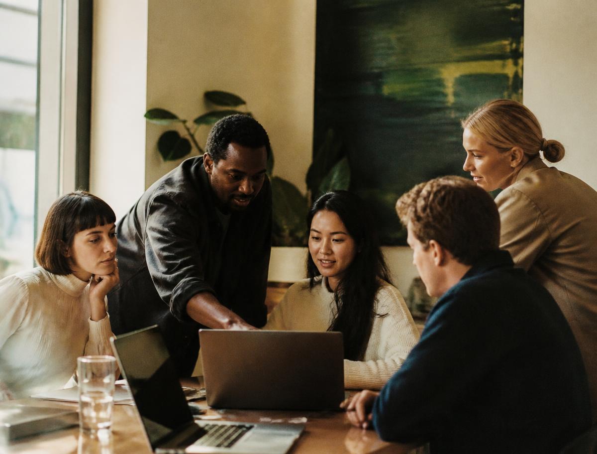 A facilitator leaning over a table, showing a laptop screen to four colleagues in a bright meeting room